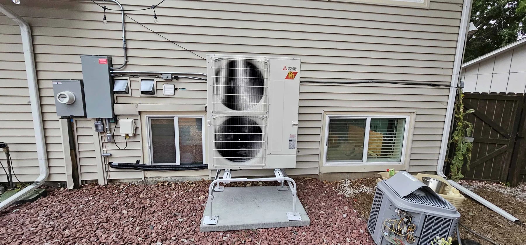 Mitsubishi two-fan outdoor unit on a stand beside beige siding, with the old AC unit next to it