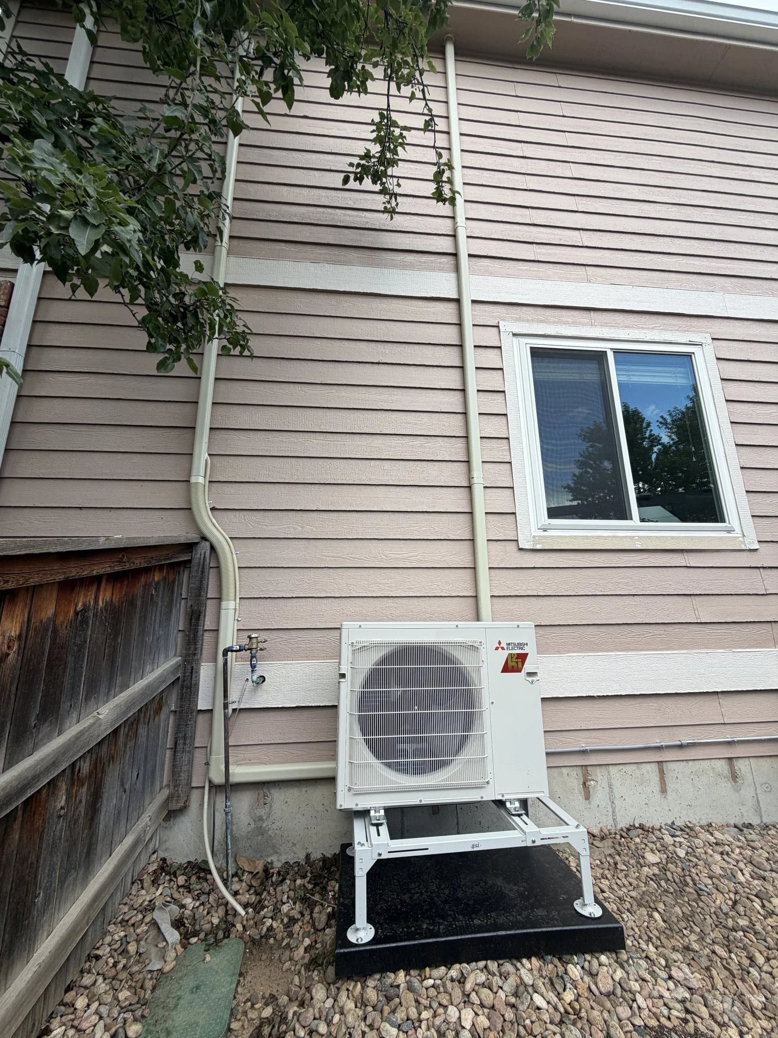 A Mitsubishi outdoor unit on a raised stand beside tan siding and a wood fence